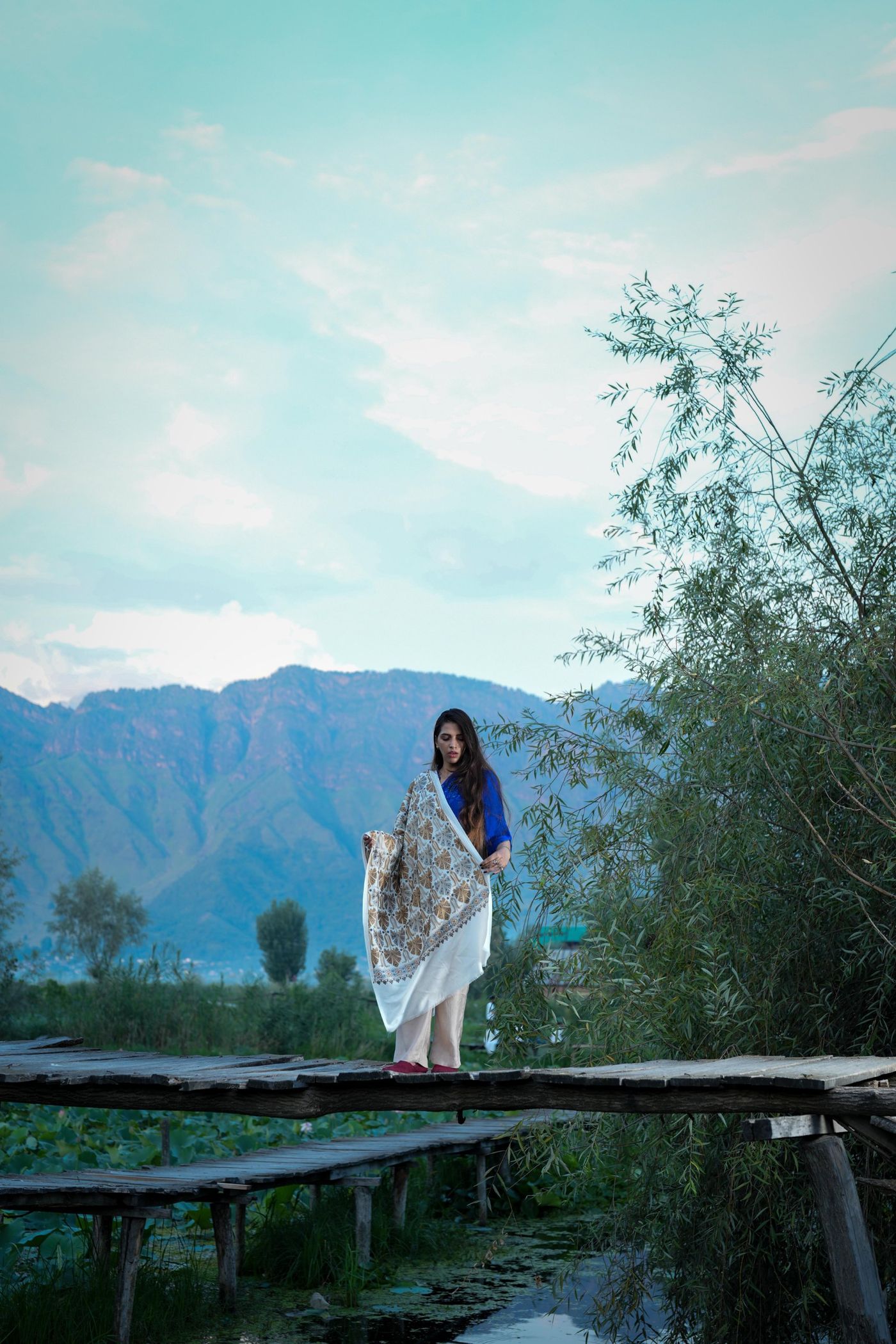 Woman wearing an embroidered ivory shawl on a wooden bridge in Kashmir, with mountains behind